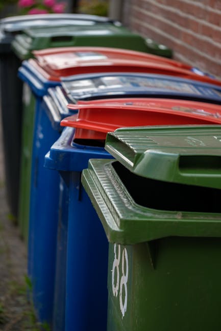 Three large black garbage bags and a rectangular cardboard box are placed on a paved sidewalk in front of a black metal fence with vertical bars. The bags are crumpled and slightly shiny, indicating t