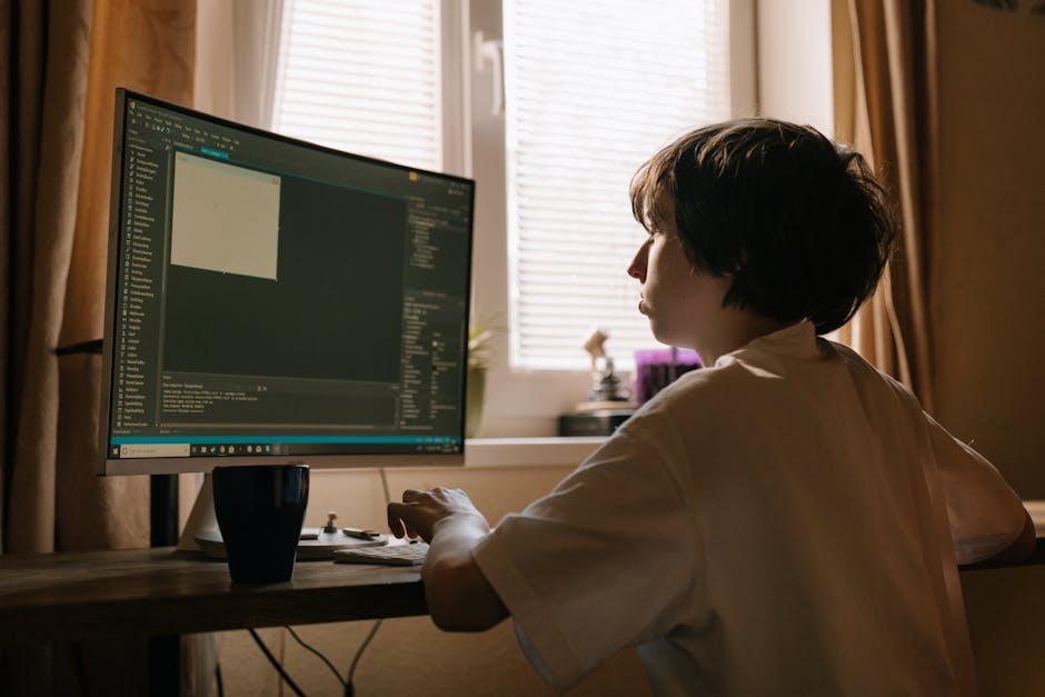 A young person with short, dark hair sits at a wooden desk inside a room with beige walls and a large window covered by partially closed white blinds. They are wearing a white shirt and are focused on